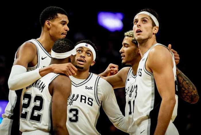 The San Antonio Spurs' starting unit (minus Devin Vassell) stands in a huddle on the court at Frost Bank Center.